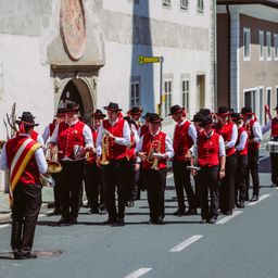 Eine Blaskapelle in roten Westen und schwarzen Hüten tritt auf einer Straße auf, mit Gebäuden und einem gelben Schild im Hintergrund.