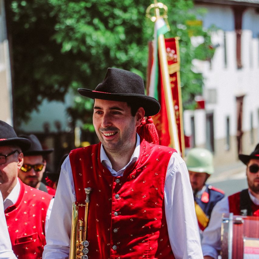 Ein Mann in einem roten Weste, schwarzem Hut und Trompete lächelt bei einer Parade mit anderen in ähnlicher Kleidung. Hinter ihm sind eine Flagge und ein Gebäude zu sehen.