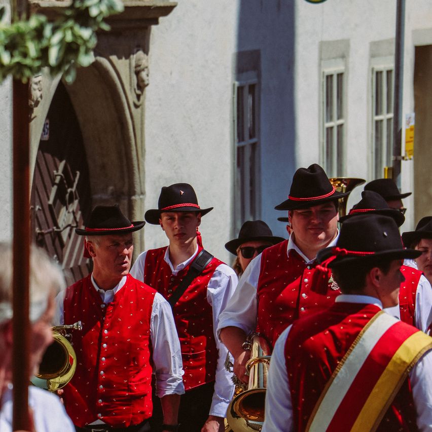 Eine Gruppe von Musikern in traditionellen roten und weißen Westen und schwarzen Hüten geht eine Straße entlang. Sie tragen Blechblasinstrumente, und ein Gebäude mit Bogenfenstern befindet sich hinter ihnen.