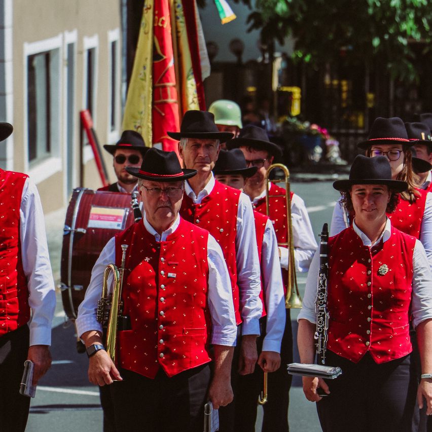 Eine Musikkapelle in roten Westen und schwarzen Hüten zieht die Straße entlang, wobei die Mitglieder Instrumente wie Trompeten und eine Trommel spielen. Dahinter sind Flaggen und ein Gebäude zu sehen.