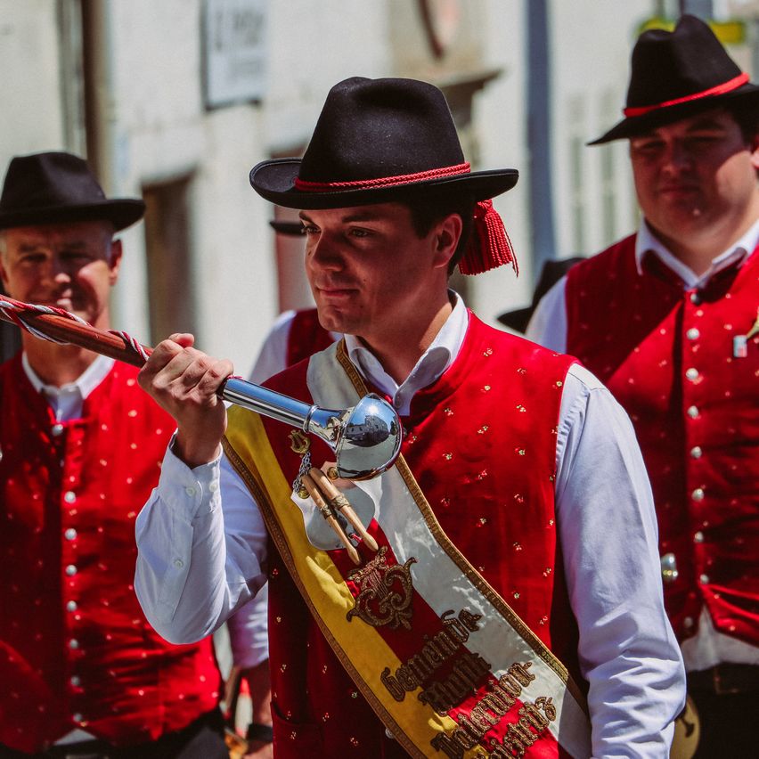 Ein Mann in einem roten Wams hält einen Stab, hinter ihm stehen zwei Männer in ähnlicher Kleidung. Sie nehmen wahrscheinlich an einer Parade oder einem Umzug teil.