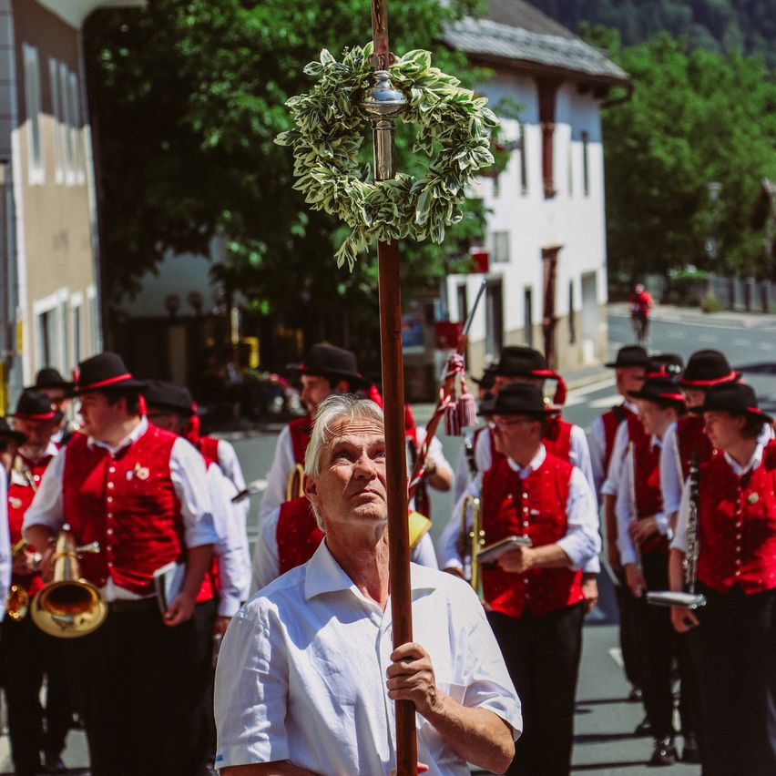 Ein älterer Mann, der einen Stab mit einem Kranz bei einer Parade hält, umgeben von Musikern in roten Westen.
