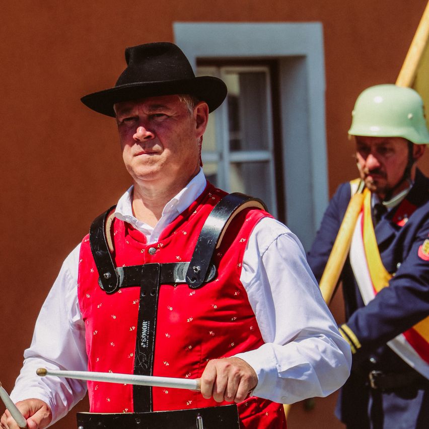 Ein Mann in einem roten Weste und schwarzem Hut hält Trommelstöcke und steht vor einem Gebäude. Ein anderer Mann mit einem grünen Helm und Uniform steht hinter ihm.