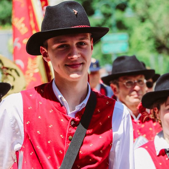 Ein Mann in einem roten Weste und schwarzem Hut lächelt, während er in einer Parade geht. Andere Menschen in roten Westen und Hüten folgen ihm. Dahinter sind verschwommene Bilder von Bäumen und Flaggen.