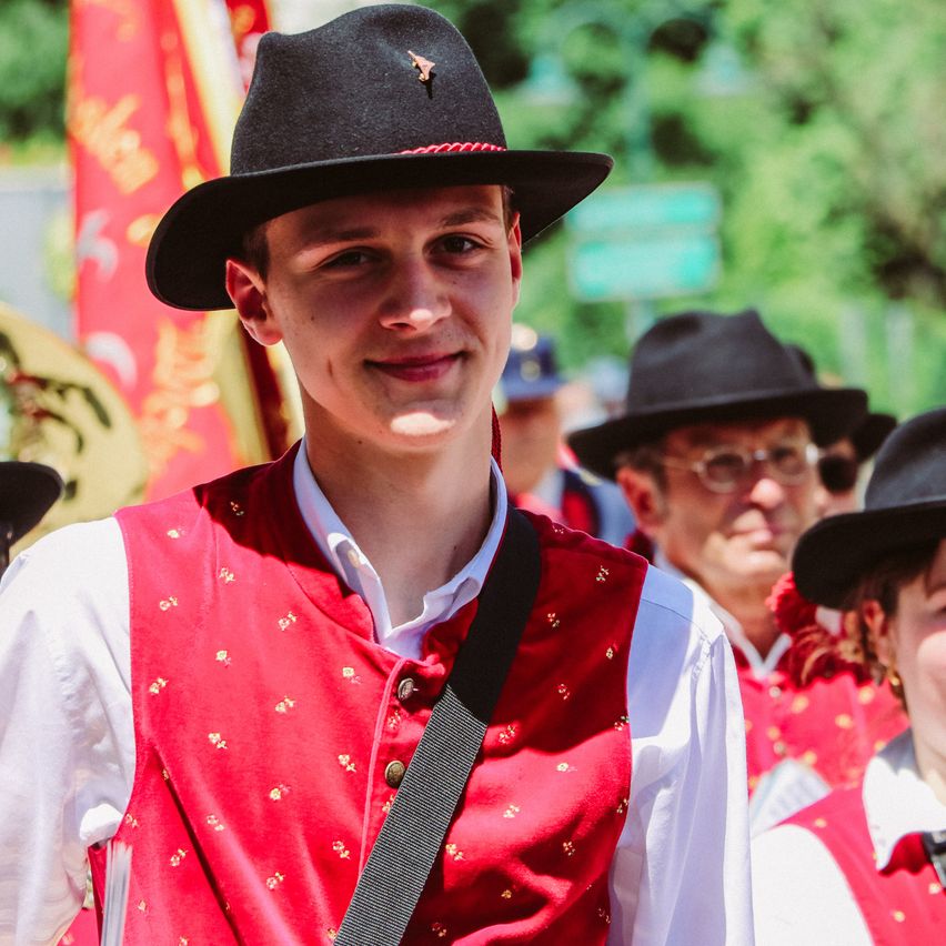 Ein Mann in einem roten Weste und schwarzem Hut lächelt, während er in einer Parade geht. Andere Menschen in roten Westen und Hüten folgen ihm. Dahinter sind verschwommene Bilder von Bäumen und Flaggen.