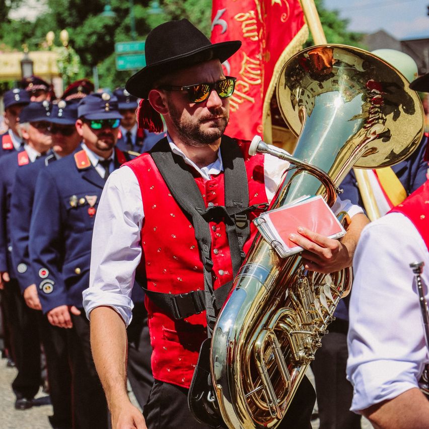 Ein Mann in einem roten Weste und Hut spielt ein großes Blechinstrument in einer Parade mit anderen Musikern in blauen Uniformen.