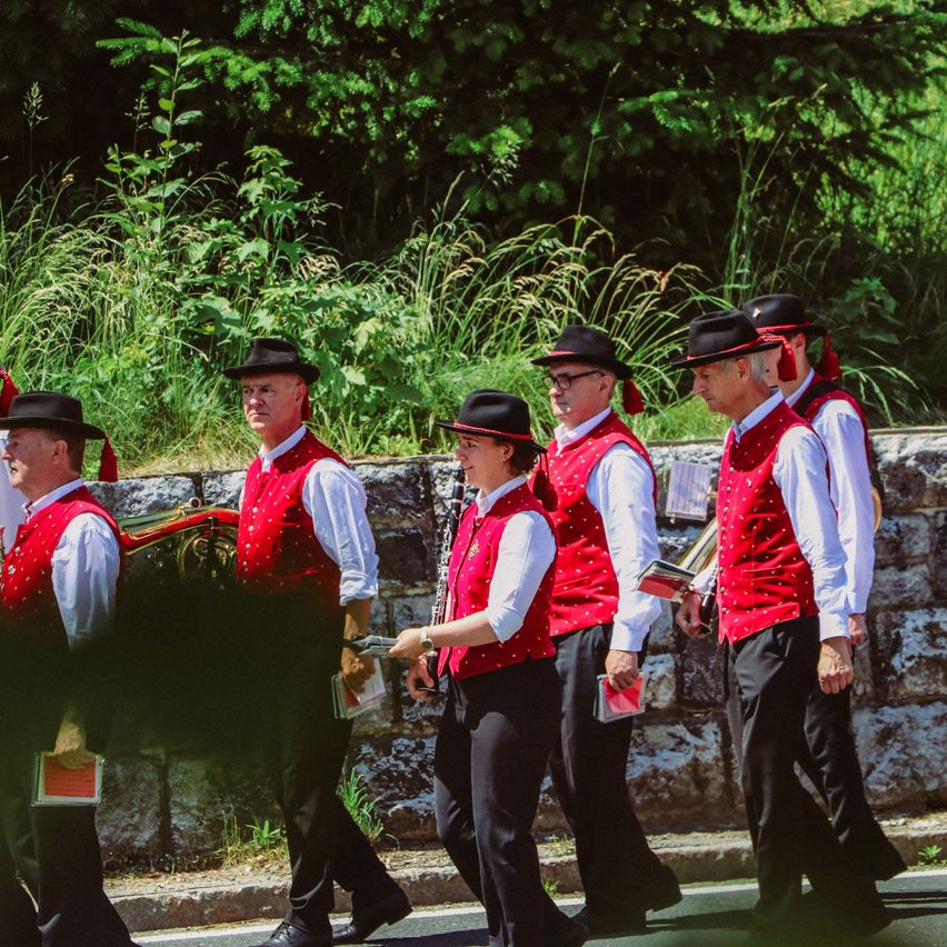 Eine Gruppe von Musikern in roten Westen und schwarzen Hüten geht eine Straße entlang, einer hält ein Buch und ein anderer trägt eine Tuba.