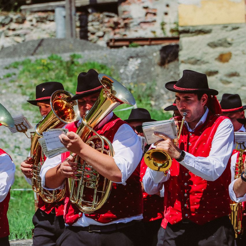 Eine Gruppe von Musikern in roten Westen und schwarzen Hüten spielt Instrumente, darunter Tubas und Trompeten, in einer Außenumgebung mit einer Steinmauer im Hintergrund.