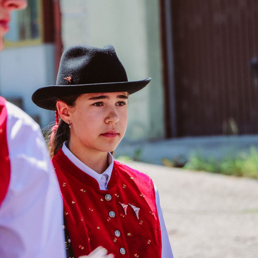 Ein Mädchen in einer roten Weste mit dekorativen Knöpfen steht vor einem Gebäude und trägt einen schwarzen Hut.