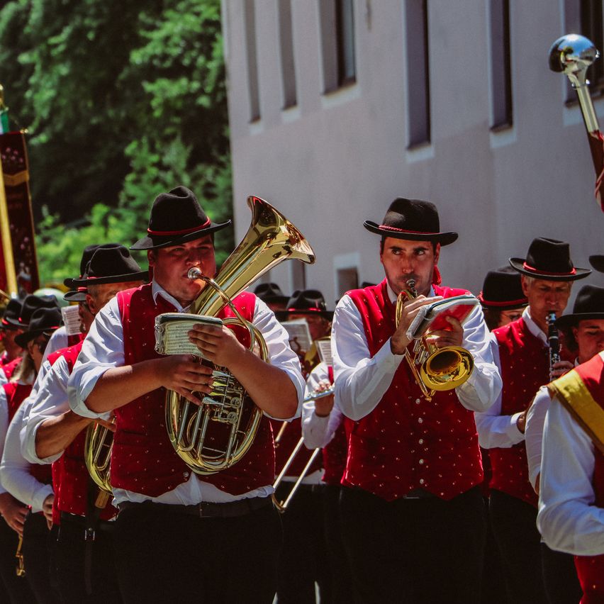 Eine Marschkapelle in roten Westen und schwarzen Hüten spielt Instrumente, darunter Trompeten und Tubas, vor einem Gebäude mit Bäumen im Hintergrund.