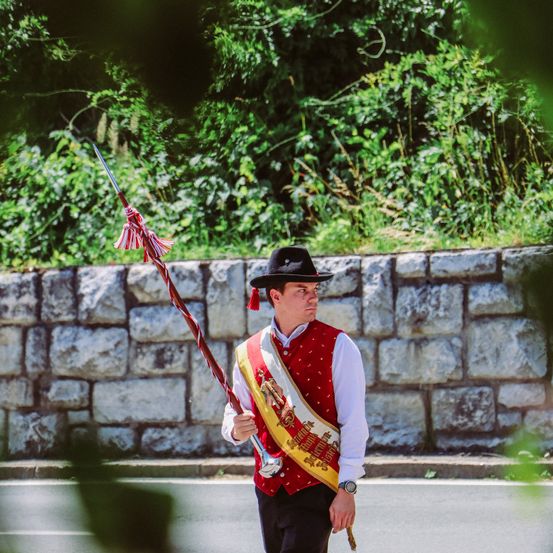 Ein Mann in traditioneller Kleidung hält einen zeremoniellen Stab und marschiert auf einer Straße, hinter ihm befindet sich eine Steinmauer.