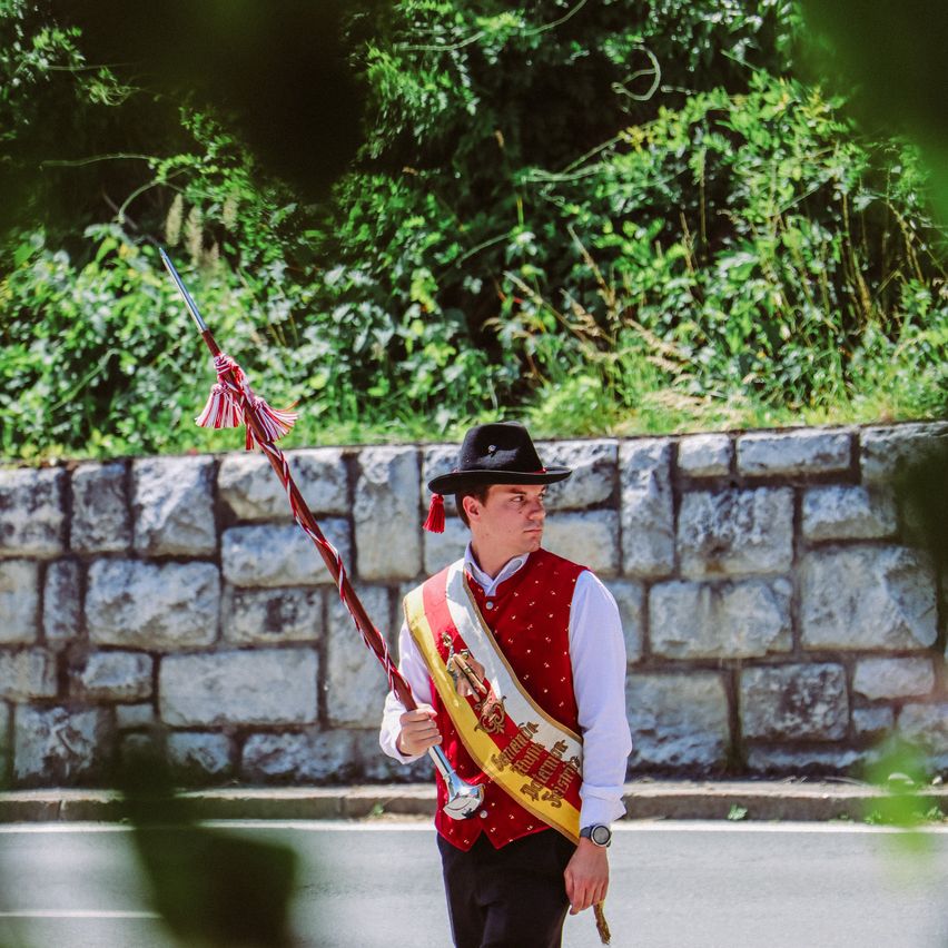 Ein Mann in traditioneller Kleidung hält einen zeremoniellen Stab und marschiert auf einer Straße, hinter ihm befindet sich eine Steinmauer.