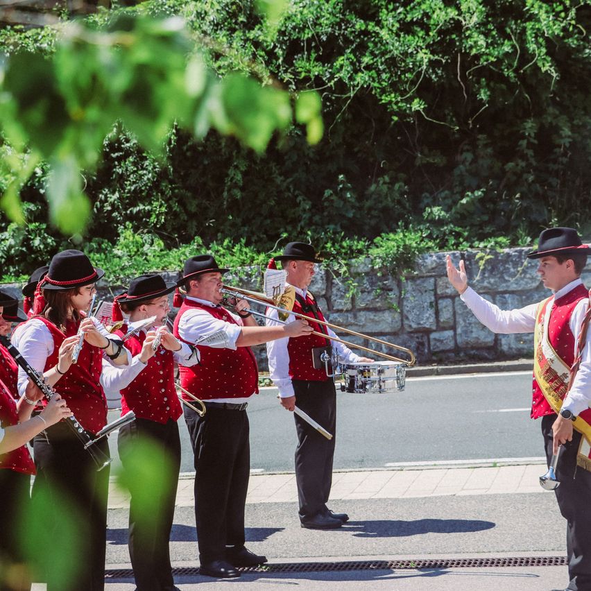 Eine Marschkapelle in roten Westen spielt Instrumente auf der Straße. Ein Mann vorne gestikuliert, und die Band steht auf dem Bürgersteig in der Nähe einer Steinmauer.
