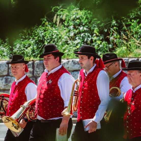 Fünf Männer in roten Westen und schwarzen Hüten, wahrscheinlich Teil einer Band, marschieren im Freien und halten Messinginstrumente in den Händen. Sie nehmen wahrscheinlich an einem Umzug teil.