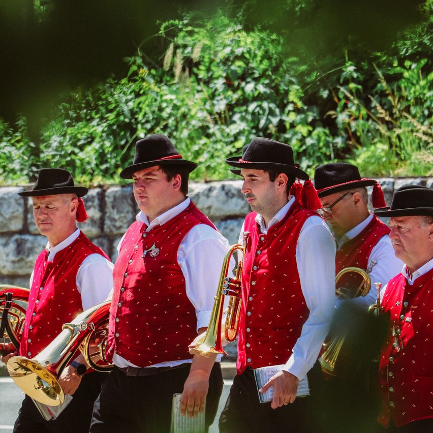 Fünf Männer in roten Westen und schwarzen Hüten, wahrscheinlich Teil einer Band, marschieren im Freien und halten Messinginstrumente in den Händen. Sie nehmen wahrscheinlich an einem Umzug teil.