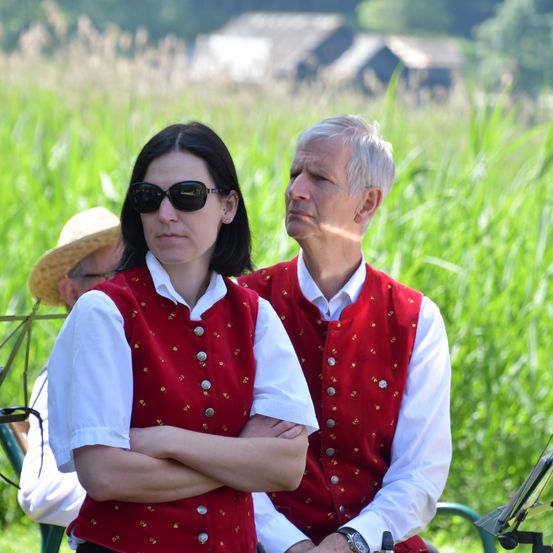 Ein Mann und eine Frau in traditioneller Kleidung stehen auf einem Feld, die Frau mit verschränkten Armen. Ein Mann mit Strohhut steht hinter ihnen.