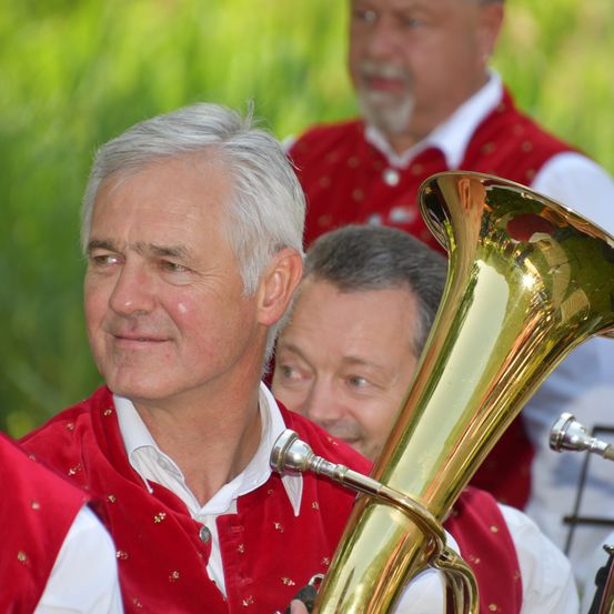 Ein Mann mit weißen Haaren spielt ein Blechblasinstrument und lächelt. Hinter ihm beobachtet ein anderer Mann. Sie tragen rote Westen.