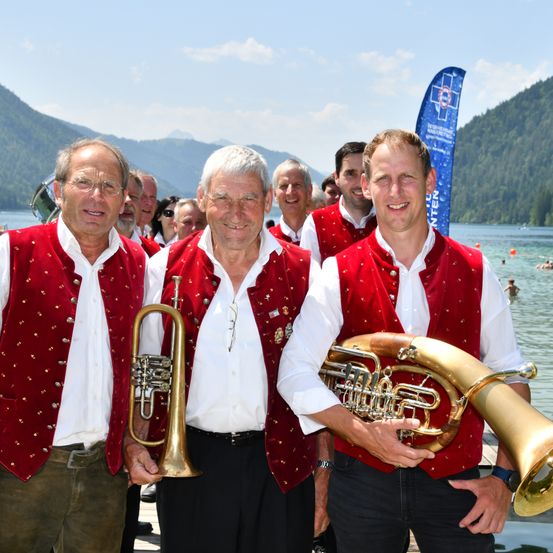 Drei Männer in roten Westen und weißen Hemden stehen auf einem Steg am See und halten Blechblasinstrumente. Hinter ihnen ist eine Gruppe von Menschen zu sehen, einige mit Sonnenbrillen. Im Hintergrund befinden sich Berge und eine blaue Flagge.