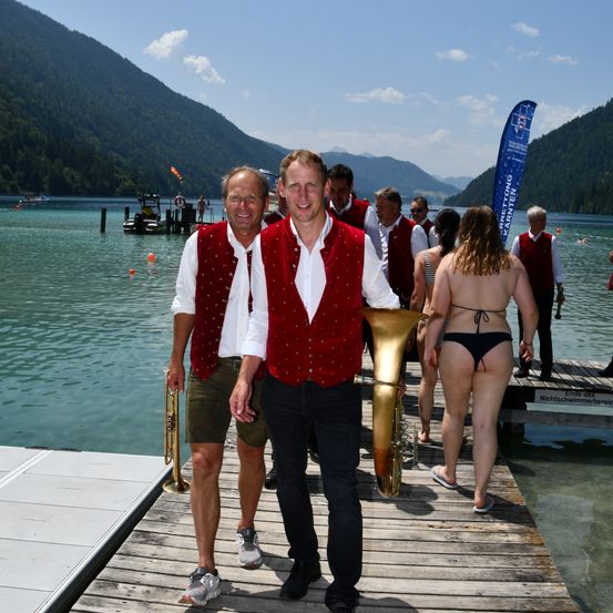 Zwei Männer in roten Westen und Shorts, Trompeten in der Hand, stehen auf einem Steg. Hinter ihnen geht eine Frau in einem Bikini zum Wasser. Im Hintergrund befinden sich Berge und eine blaue Fahne.
