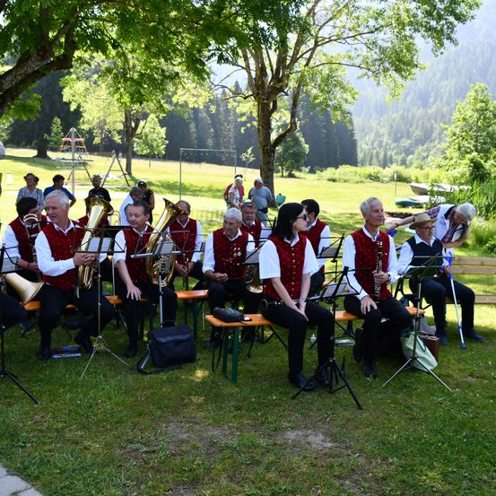 Eine Gruppe von Musikern spielt auf einer Wiese mit Bäumen und Bergen im Hintergrund. Sie sind in roten Westen und schwarzen Hosen gekleidet.