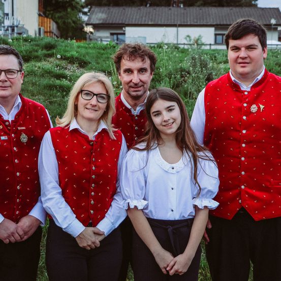 Eine Familie mit fünf Personen steht auf einem Rasen und trägt rote Westen und schwarze Hosen. Sie posieren für ein Foto mit einem Haus und Bäumen im Hintergrund.