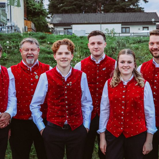 Eine Gruppe von Menschen in roten Westen und schwarzen Hosen steht auf einem Feld mit einem Haus im Hintergrund. Die Westen haben dekorative Muster.