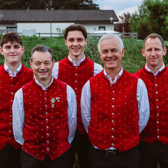 Fünf Männer in roten Westen und weißen Hemden posieren für ein Foto im Freien. Sie stehen in einem Feld mit grünem Gras und Bäumen im Hintergrund. Der Mann rechts trägt einen Gürtel.