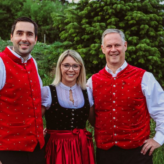Drei Menschen in traditioneller Kleidung, lächelnd, stehen vor einer großen immergrünen Tanne. Die Frau trägt einen roten Rock und die Männer rote Westen.