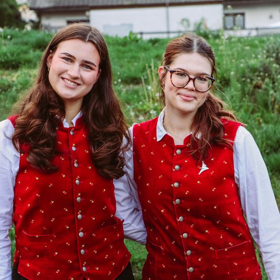 Zwei Frauen stehen lächelnd in einem Feld mit einem Haus im Hintergrund. Sie tragen passende rote Westen mit dekorativen Knöpfen.