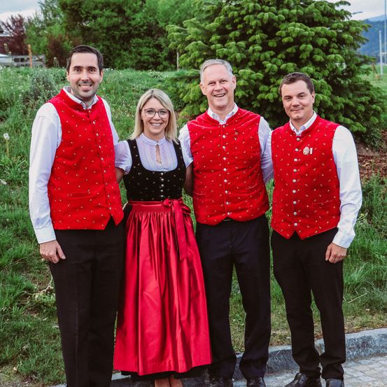 Vier Menschen stehen eng nebeneinander auf dem Bürgersteig. Drei Männer tragen rote Westen mit Silberknöpfen. Eine Frau in der Mitte trägt ein rotes Kleid. Dahinter befindet sich ein Grasfeld mit Bäumen.