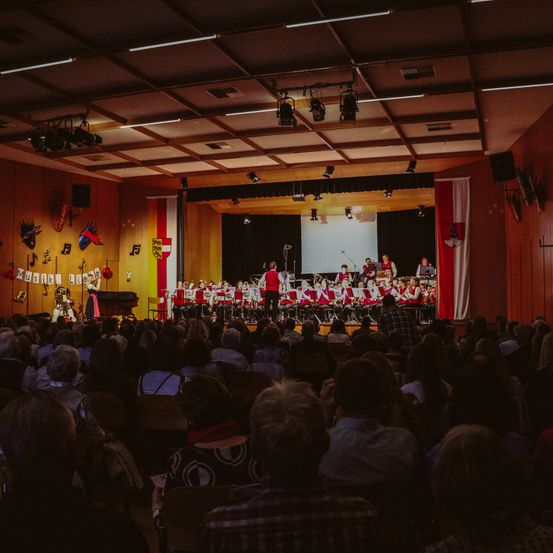 Ein Chor tritt auf der Bühne mit einem Projektor im Hintergrund auf. Das Publikum, in Reihen sitzend, beobachtet aufmerksam. Die Bühne ist mit rot-weißen Bannern geschmückt.