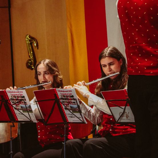 Zwei junge Frauen spielen Flöten im Sitzen und tragen rote Westen. Hinter ihnen ist ein rot-gelber Hintergrund mit einer goldenen Musiknote. Sie lesen von Notenständern.