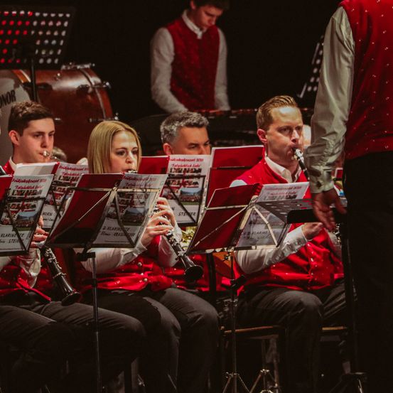 Eine Gruppe von Musikern in roten Westen tritt auf der Bühne auf. Eine Frau spielt Klarinette, während ein Mann Trompete spielt. Hinter ihnen sind weitere Musiker zu sehen.