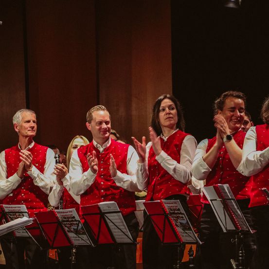 Eine Gruppe von Musikern, die rote Westen und weiße lange Ärmel tragen, tritt auf der Bühne auf, klatscht und lächelt. Die Bühne hat einen dunklen Hintergrund und einige Notenständer mit Notenblättern.