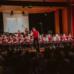Ein Blasorchester spielt auf der Bühne mit einem Dirigenten. Das Publikum sitzt davor und eine Leinwand zeigt ein Bild hinter der Band.