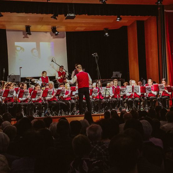 Ein Blasorchester spielt auf der Bühne mit einem Dirigenten. Das Publikum sitzt davor und eine Leinwand zeigt ein Bild hinter der Band.