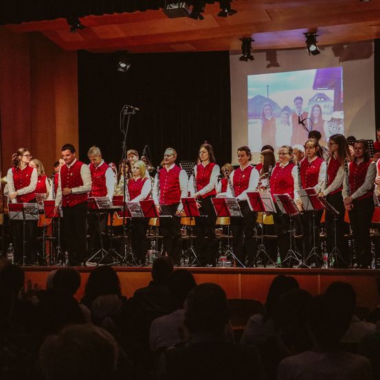 Ein Chor in roten Westen und schwarzen Hosen tritt mit Musikinstrumenten auf der Bühne auf. Ein Bildschirm hinter ihnen zeigt ein Gruppenfoto. Das Publikum sitzt vor der Bühne.