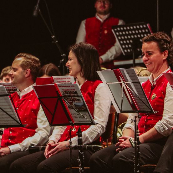 Eine Gruppe von Musikern in roten Westen und weißen Hemden sitzen auf der Bühne in Stühlen, lächeln und haben Notenständer und Instrumente vor sich.