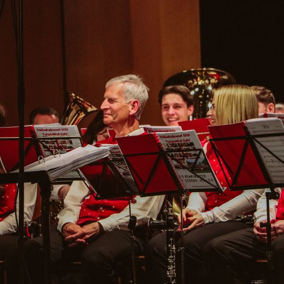 Ein Orchester sitzt auf der Bühne und bereitet sich auf einen Auftritt vor. Die vorderste Reihe in roten Westen hält Notenblätter. Dahinter stehen ein Posaunenspieler und andere, die zuschauen.