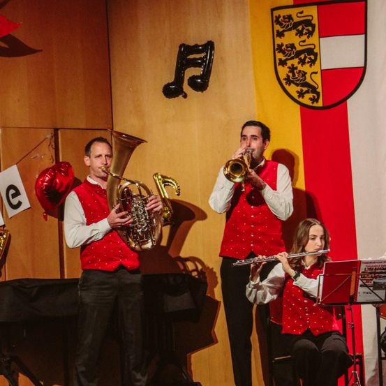 Drei Musiker stehen auf der Bühne; zwei Männer spielen Blasinstrumente, während eine Frau Flöte spielt. Hinter ihnen ist ein Banner mit einem Wappen und einer musikalischen Note.