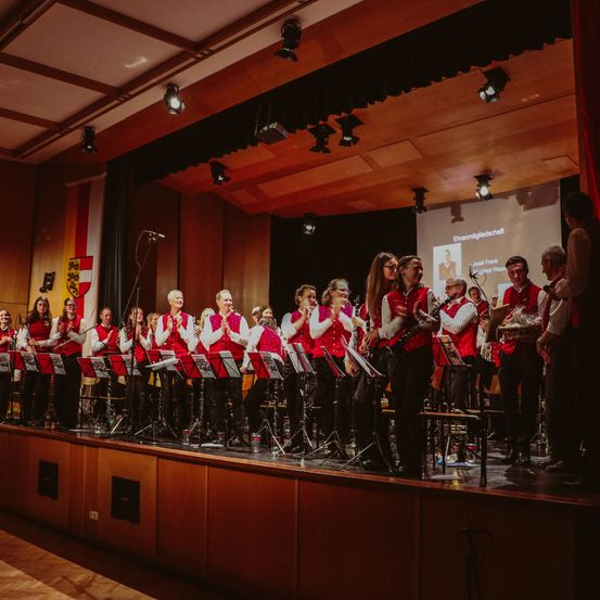 Eine Gruppe von Musikern in roten Westen und schwarzen Hosen spielt auf der Bühne, mit Instrumenten wie Klarinetten und Trompeten. Eine Projektionsleinwand zeigt das Foto und den Text einer Person, und ein Mikrofon steht auf der linken Seite.