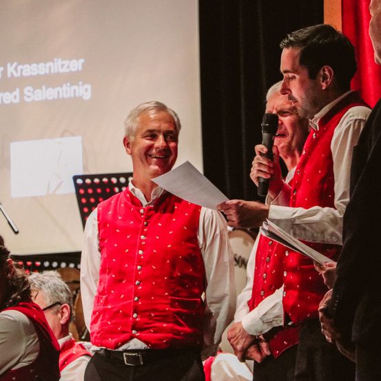 Eine Gruppe von Männern in roten Westen mit weißen Hemden und schwarzen Hosen singt und hält ein Mikrofon und Papiere in einem schwach beleuchteten Raum.