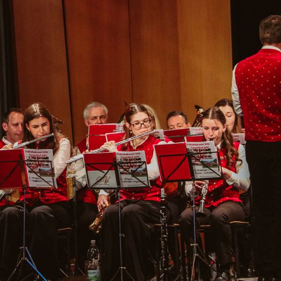 Eine Gruppe von Musikern in roten Westen spielt Instrumente auf der Bühne. Sie haben Notenständer vor sich mit Notenblättern. Ein Mann in einem roten West steht hinter ihnen.