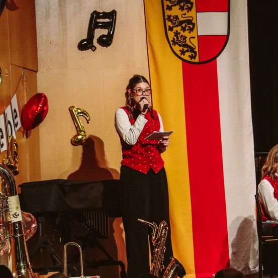 Eine Frau steht auf der Bühne, hält ein Mikrofon und Papier in der Hand, mit einer Flagge, Instrumenten und einem roten Luftballon im Hintergrund.