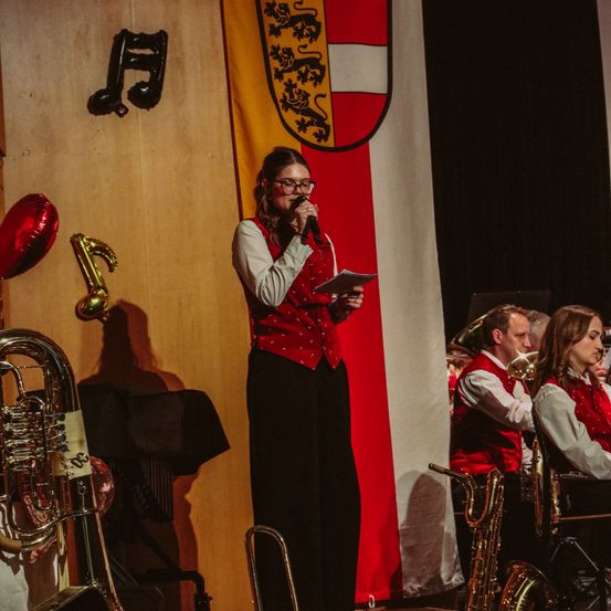 Eine Frau mit Brille und Mikrofon steht auf einer Bühne mit einer Fahne dahinter. Sie hält ein Papier und liest daraus vor. Musikinstrumente sind links, und eine Band sitzt hinter ihr.