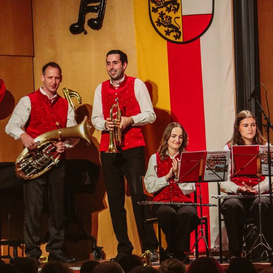 Eine Band mit vier Mitgliedern spielt auf der Bühne. Drei Männer und eine Frau stehen und sitzen. Sie tragen rote Westen. Sie haben Musikinstrumente vor sich. Hinter ihnen ist eine Flagge mit einem Löwenwappen.