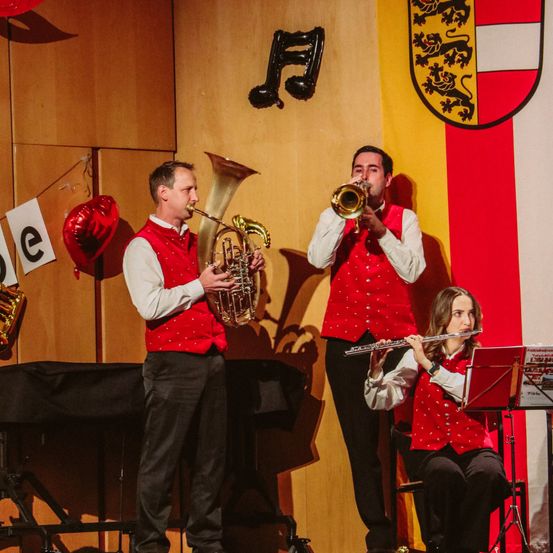 Drei Musiker treten auf der Bühne auf. Zwei Männer spielen Trompeten, ein Mann spielt Tuba und eine Frau Flöte. Ein Wappen hängt hinter ihnen.