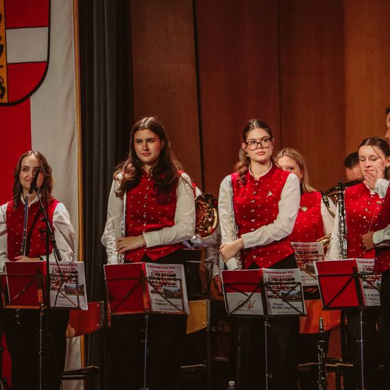 Eine Gruppe junger Frauen in roten Westen und Brillen spielt mit Musikinstrumenten auf der Bühne. Ein Fahnen im Hintergrund ist sichtbar.