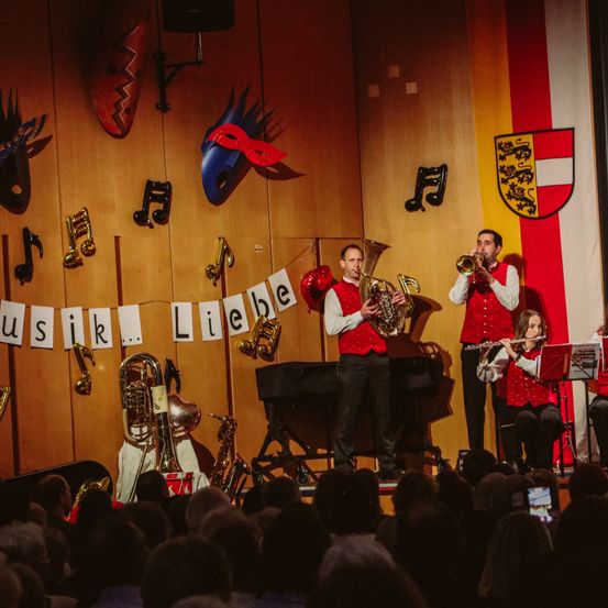 Eine Brass Band spielt auf der Bühne, mit einer Frau, die Flöte spielt, zwei Männern, die Trompeten spielen, und verschiedenen Musikinstrumenten an der Wand. Das Publikum sitzt vor ihnen.
