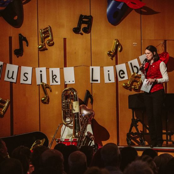 Eine Frau spricht in ein Mikrofon auf einer Bühne mit Musikinstrumenten und einem Publikum. Musiknoten sind an der Wand hinter ihr angebracht.
